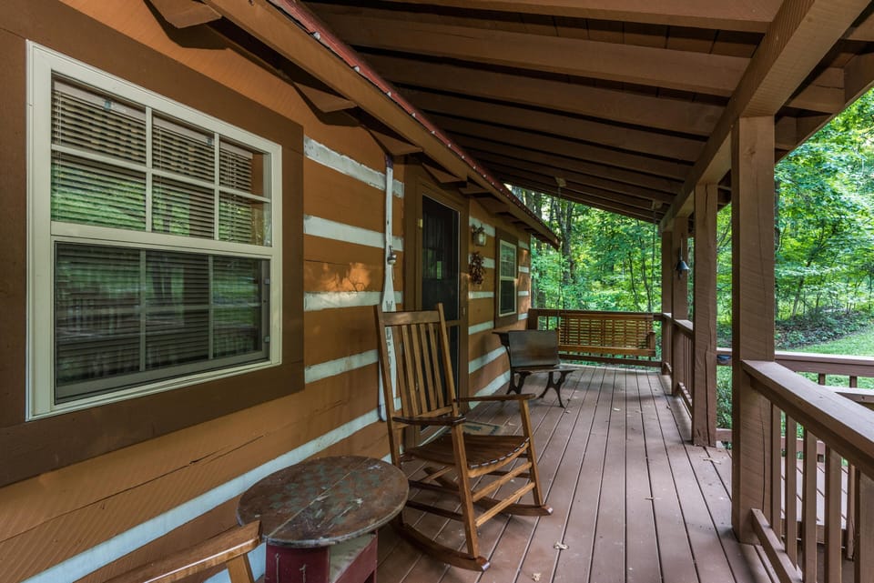 Covered front porch with wooden rocking chair, metal bench, and brown plank flooring surrounded by trees.