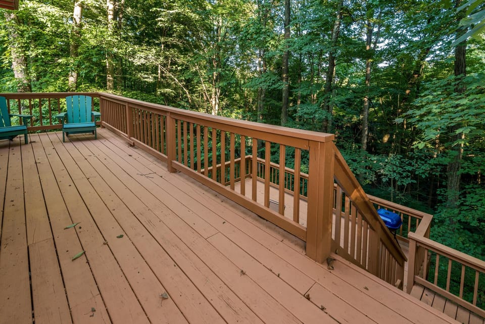 Expansive wooden deck with brown railing, two teal Adirondack chairs, and surrounding lush green forest.