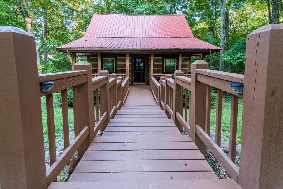 Wide wooden ramp with railings leads to the log cabin’s front porch, surrounded by dense green trees.