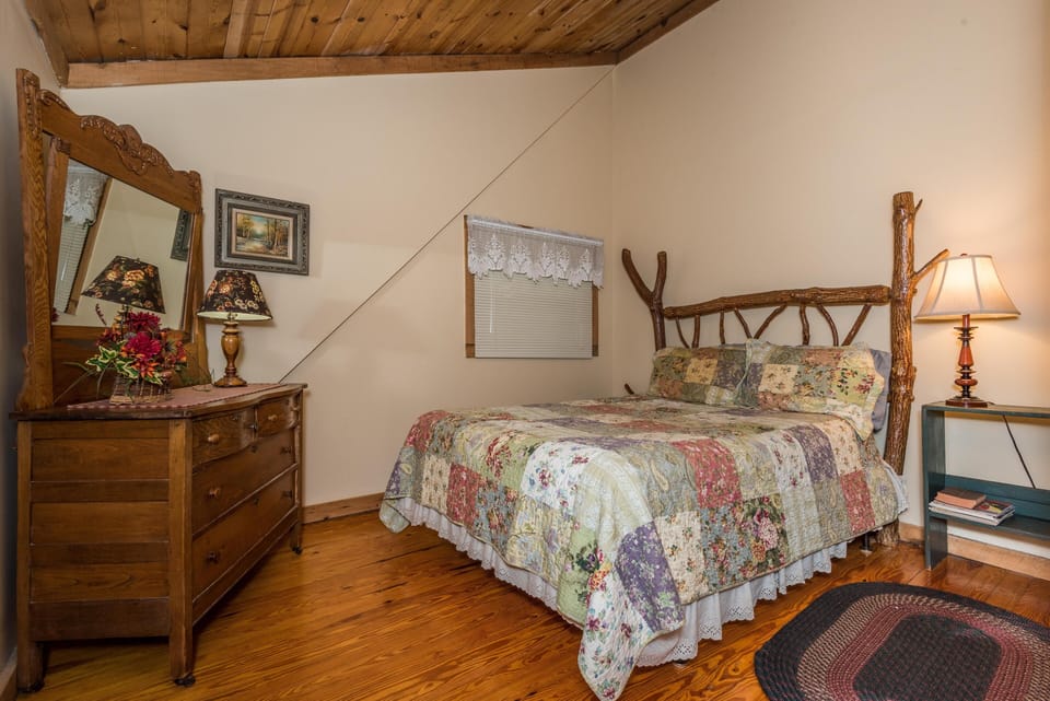 Rustic bedroom with a handcrafted log bed frame, floral patchwork quilt, wooden dresser, and lace-trimmed window curtain.