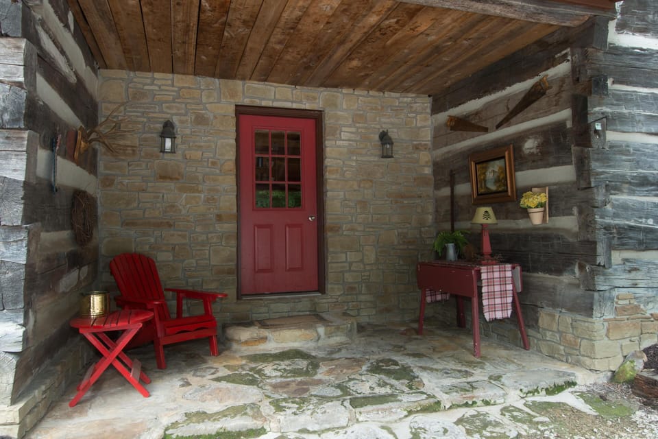Covered cabin porch with flagstone floor, weathered log walls, red Adirondack chair, and matching side table.