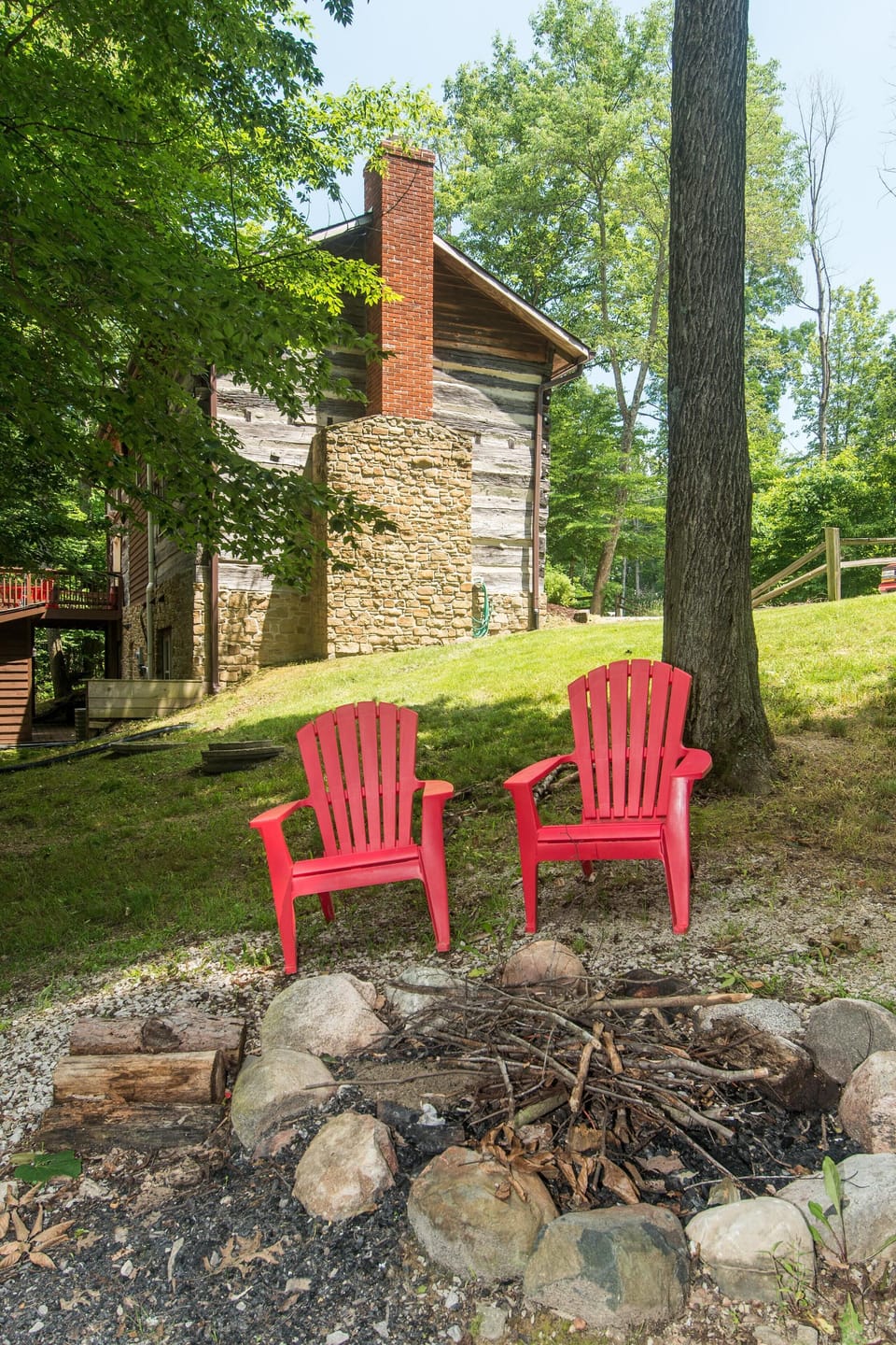 Fire pit encircled by large stones, with two red Adirondack chairs on gravel and grass near a log cabin.