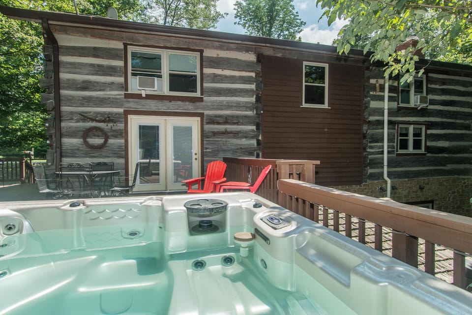 Hot tub on wooden deck overlooks log cabin exterior with stone foundation and red Adirondack chairs.