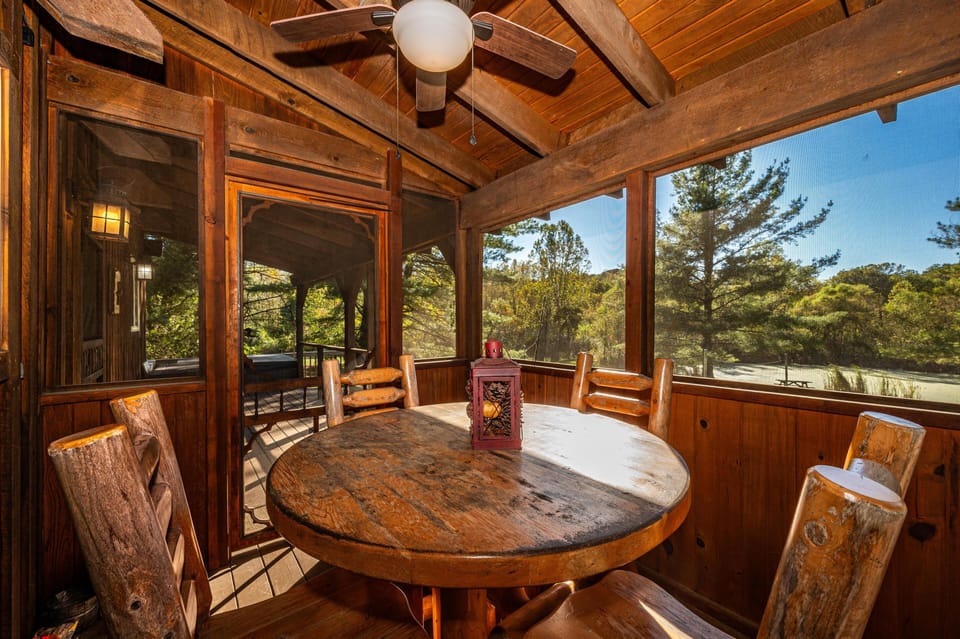 Screened porch dining area with round wood table, log chairs, ceiling fan, and views of surrounding pine trees.