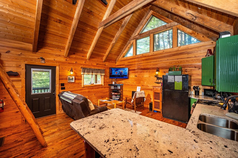 Granite countertop island overlooks living space with leather recliners, green cabinets, and vaulted pine ceiling.