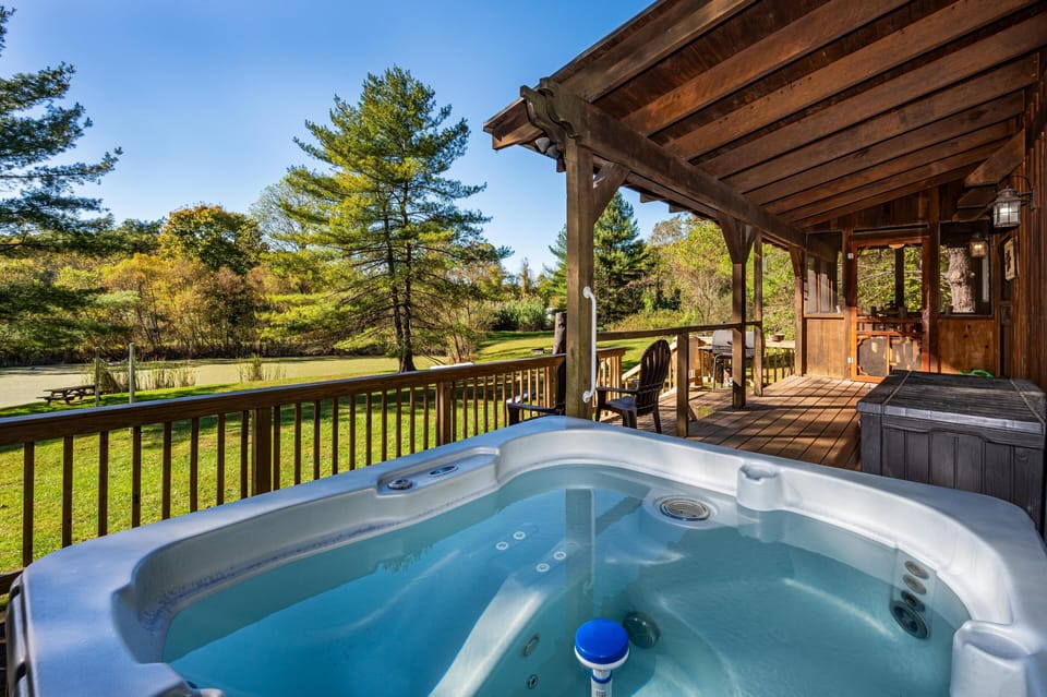 Outdoor hot tub on wooden deck under a covered porch, overlooking a grassy yard and pond with picnic table.
