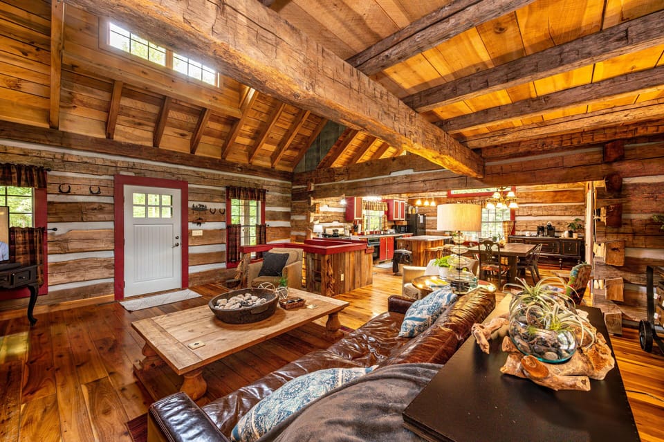 Open living area with exposed log beams, wide plank floors, leather sofa, and red-accented kitchen cabinetry.