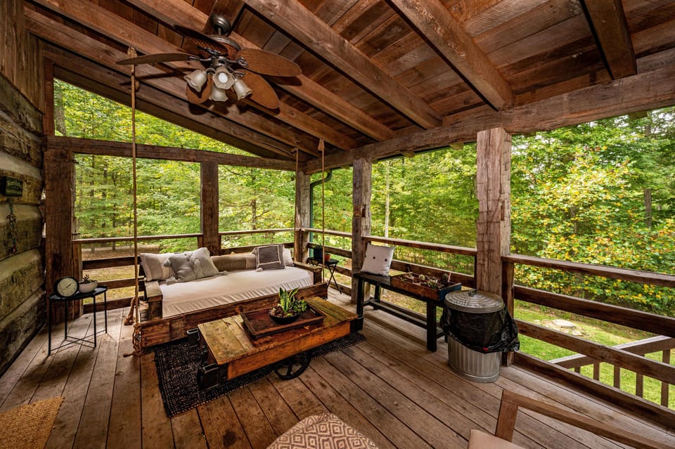 Covered porch with a hanging daybed, wooden coffee table on wheels, and views of surrounding forest greenery.