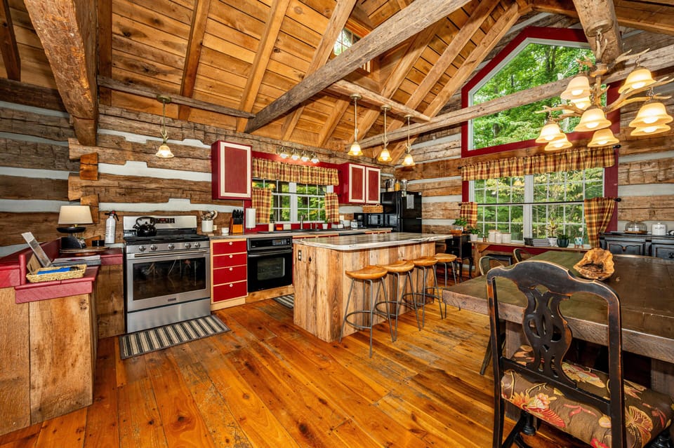 Log cabin kitchen with red cabinetry, marble-topped island, exposed beams, and plaid curtains framing large windows.
