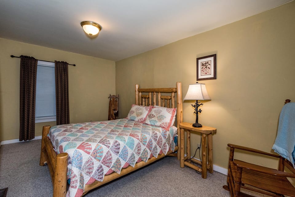 Log-framed bed with a colorful patchwork quilt, matching wood nightstand, and rustic chair by beige walls.
