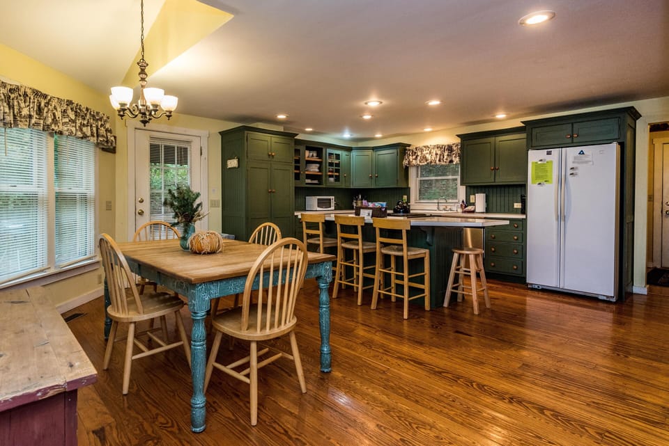 Dining area with wooden table, spindle-back chairs, and a bench beneath windows with patterned valances.