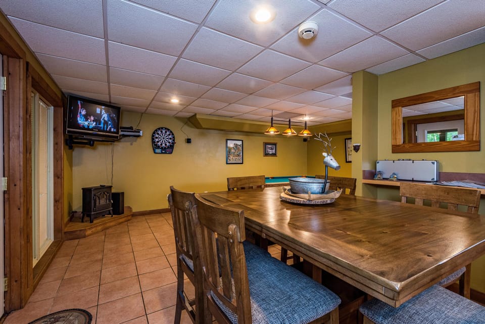 Game room with wooden dining table, dartboard, wall-mounted TV, and tile flooring.