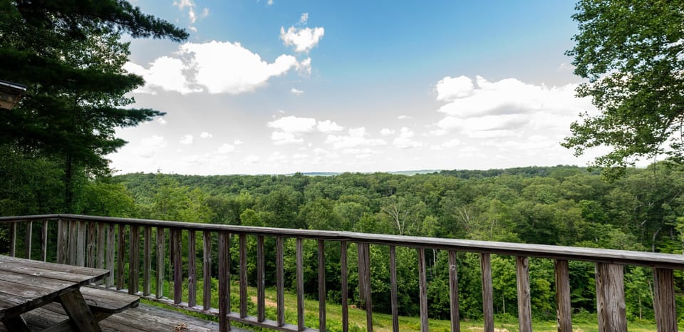 A weathered wood picnic table sits on the deck beside Adirondack chairs and a charcoal grill.