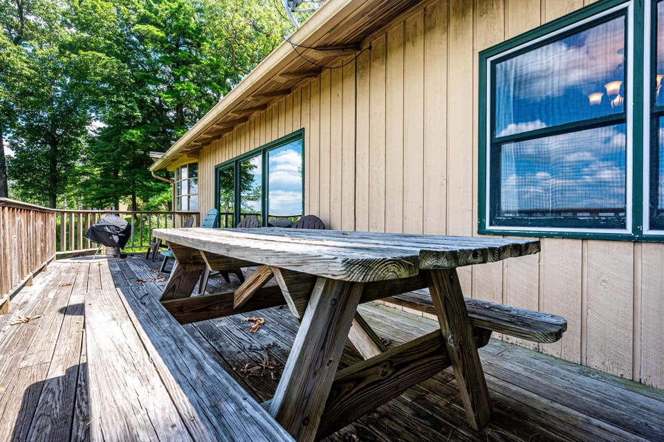 Weathered wood picnic table and bench on the deck with Adirondack chairs and a charcoal grill nearby.