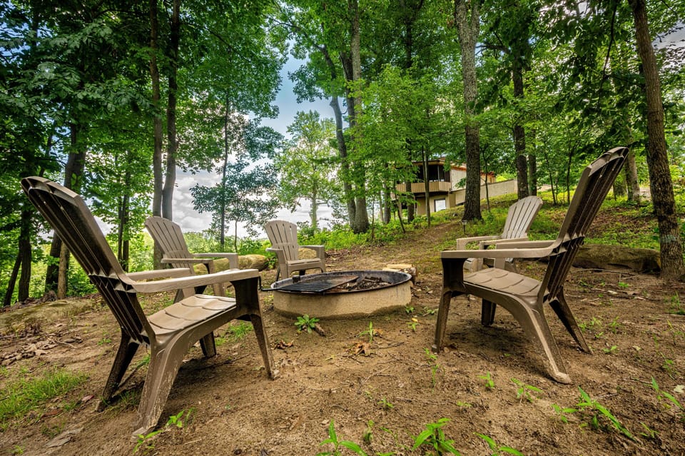 Outdoor fire pit encircled by five molded plastic chairs set on dirt ground beneath tall leafy trees.