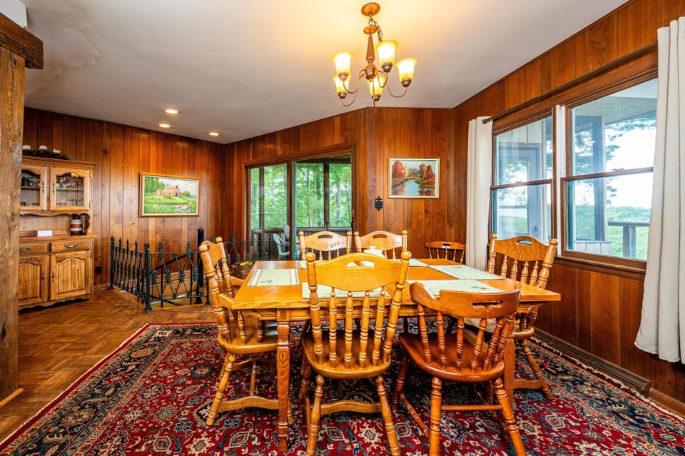 Dining room with honey-toned wood table and spindle-back chairs on a red patterned rug by large windows.