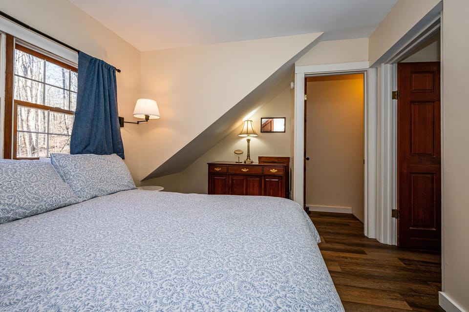 Soft blue patterned bedding covers the bed beneath a window with denim curtains, beside a wall-mounted lamp.