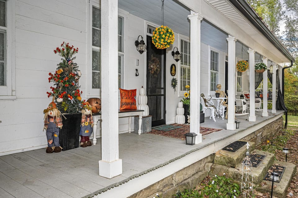 Wide front porch with white columns, hanging flower baskets, seasonal fall décor, and white metal bistro set.