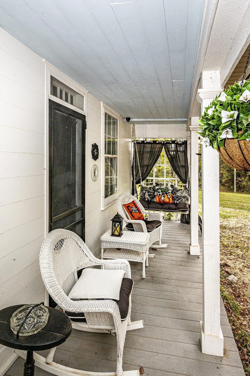 Front porch with white wicker rocking chairs, side tables, a hanging bench with black cushions, and seasonal pillows.