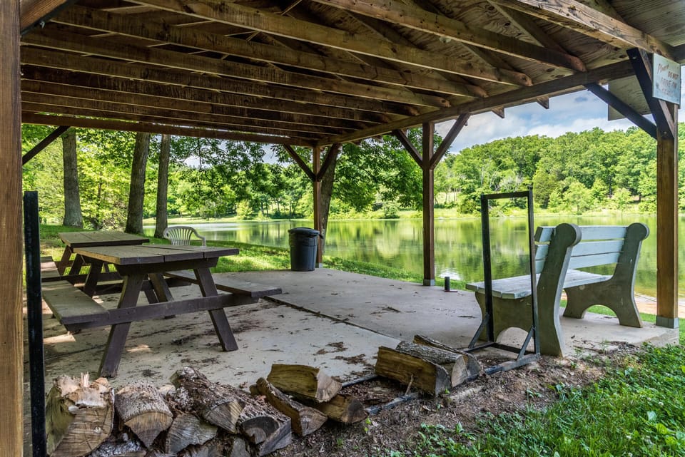 Covered lakeside pavilion with brown wooden picnic table, green bench, and stacked firewood nearby.