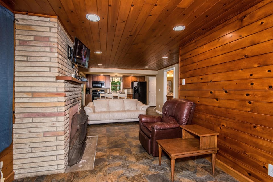 Living room with a stone fireplace, mounted TV, leather recliner, cream sofa, and knotty pine wood walls.