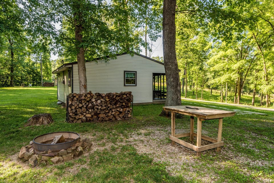 Outdoor fire pit surrounded by stones, stacked firewood, and a rustic wooden prep table under tall leafy trees.