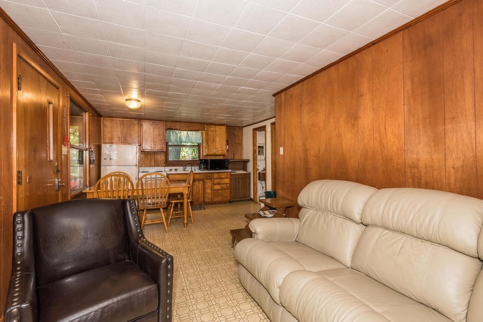 Living area with cream leather sofa and dark armchair faces wood-paneled walls and vintage linoleum flooring.