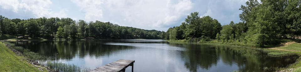 View across the calm lake with a weathered wooden dock extending over the water, surrounded by dense green trees.