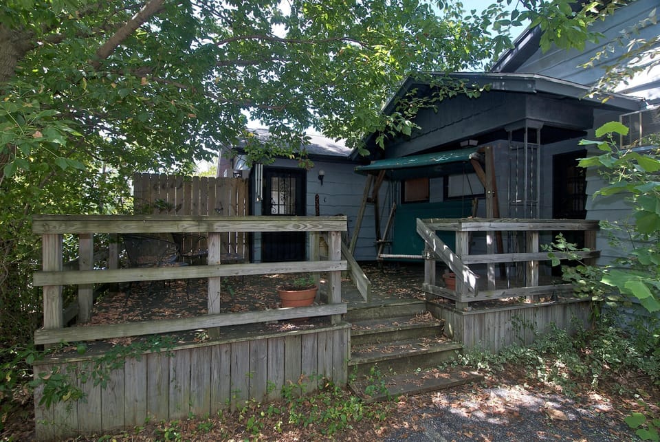 Shaded wooden deck with railing, green canopy porch swing, and black metal patio chairs surrounded by trees.