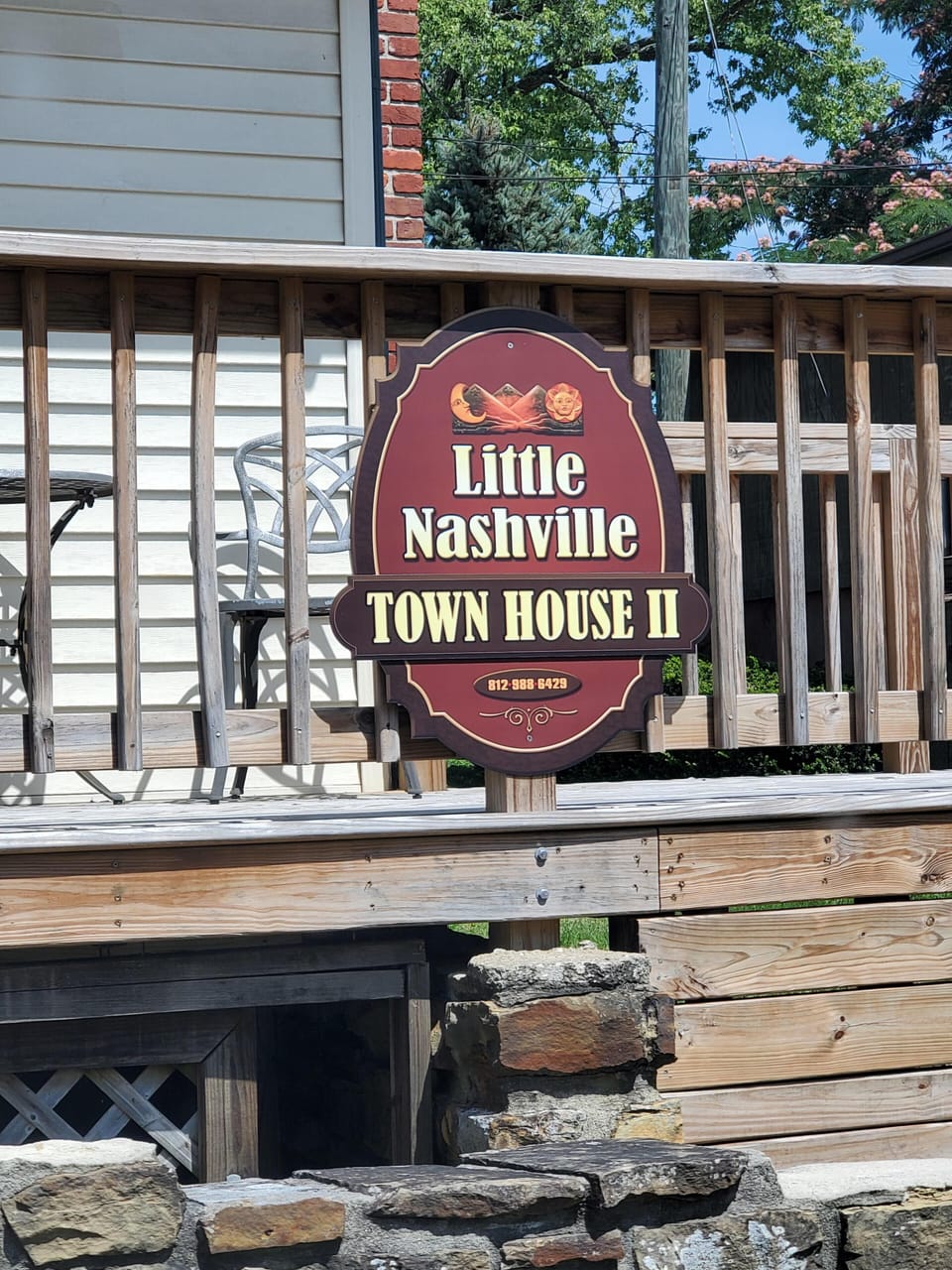 Wooden deck railing with a maroon and cream Little Nashville Town House II sign, metal chairs, and green trees beyond.