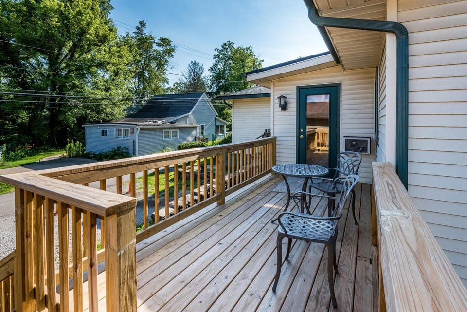 Sunlit wooden deck with a black metal table and two chairs, overlooking green trees and neighboring homes.