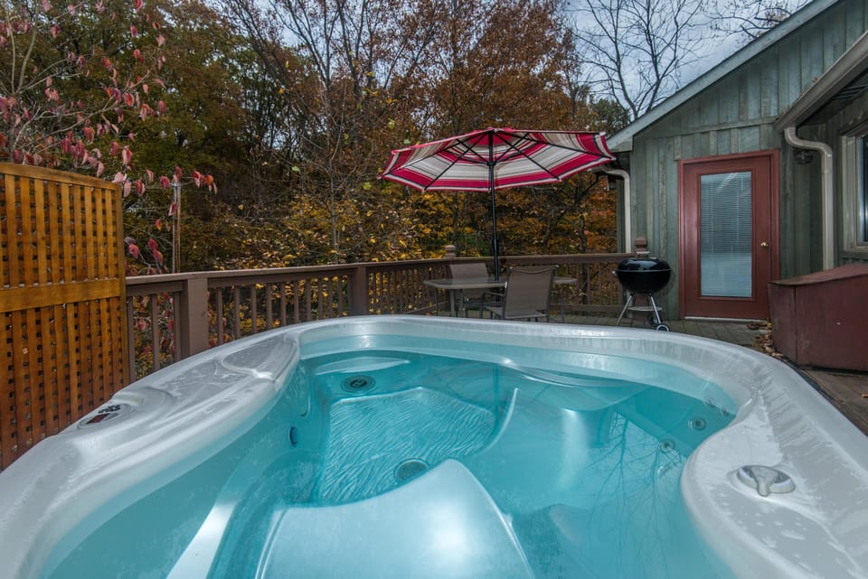 Outdoor deck with filled hot tub, glass-top table under red-striped umbrella, charcoal grill, and wood privacy screen.