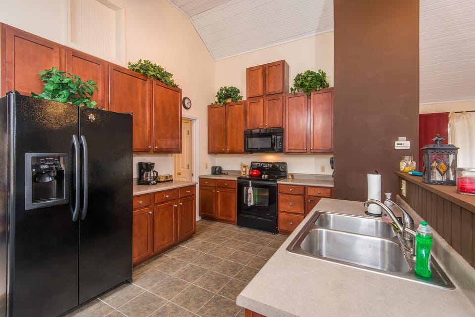 Kitchen features dark appliances, wood cabinets topped with greenery, beige countertops, and tile flooring under vaulted ceiling.