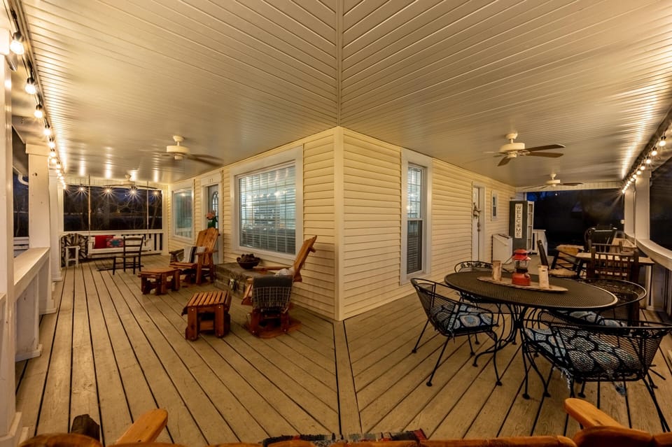 Wraparound porch with wood plank flooring, hanging string lights, ceiling fans, metal dining set, and rustic wooden chairs.