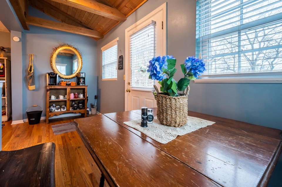 Natural light fills the dining nook with blue walls, wooden table, woven basket centerpiece, and coffee station below a round mirror.