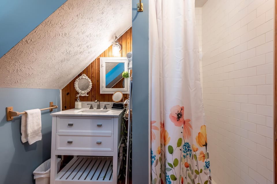 Sloped ceiling frames a white vanity with marble countertop beside a shower with floral curtain and subway tile surround.