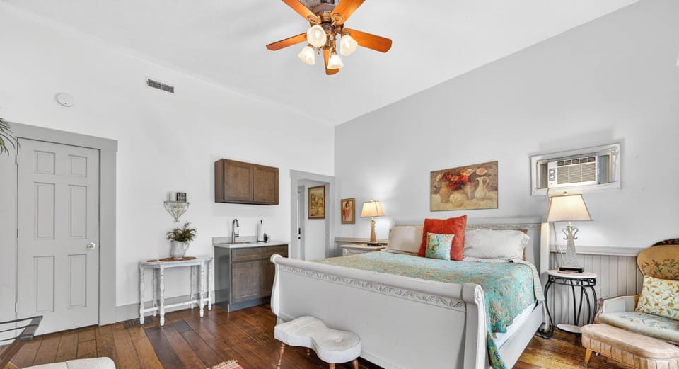 White sleigh bed with floral quilt sits beside a vintage armchair and hardwood flooring in the bedroom.