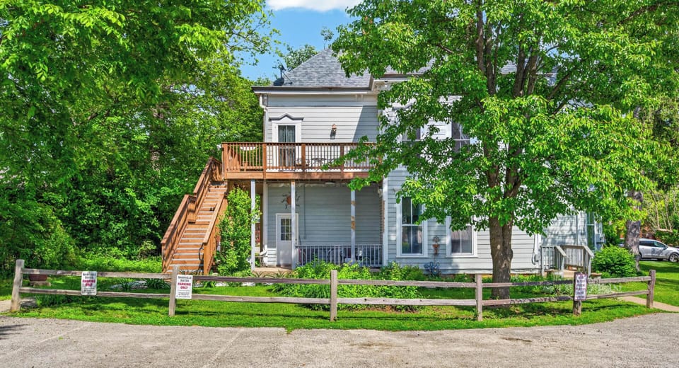 Exterior view of white Victorian-style apartment with wooden staircase, deck, and lush green trees surrounding parking area.