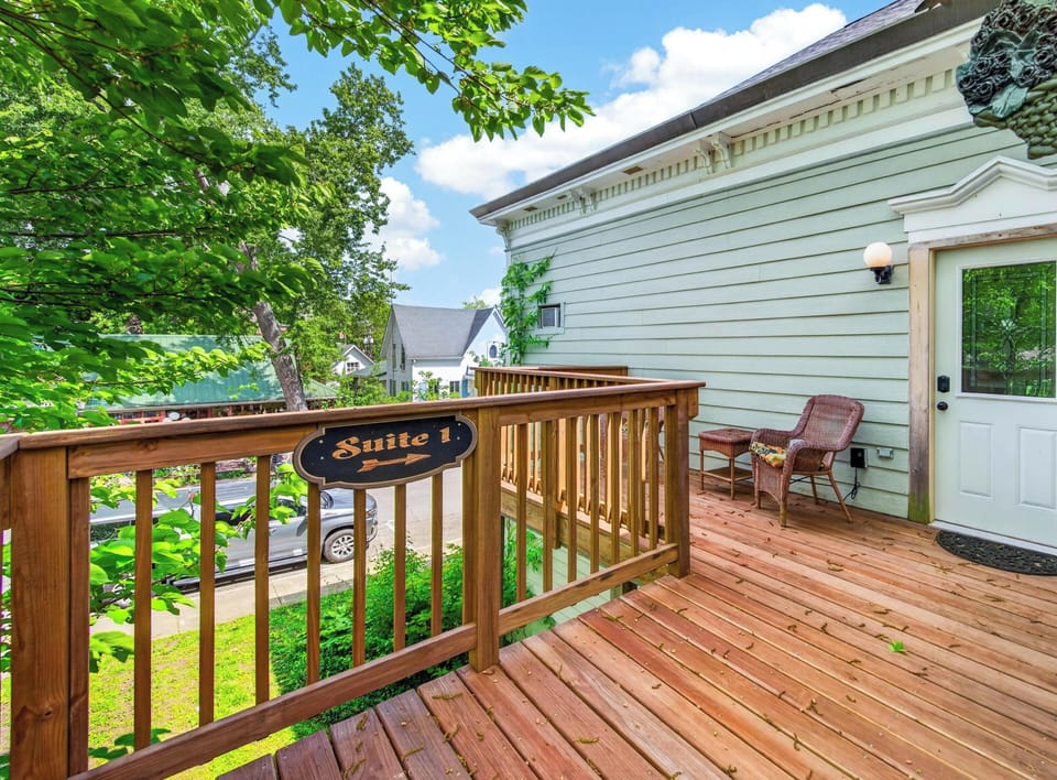 Wood-plank balcony with wicker seating overlooks leafy trees and neighboring homes under a bright blue sky.