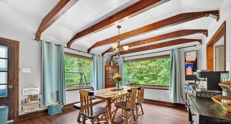 Rustic wood dining table and chairs sit beneath exposed ceiling beams by large corner windows with blue curtains.