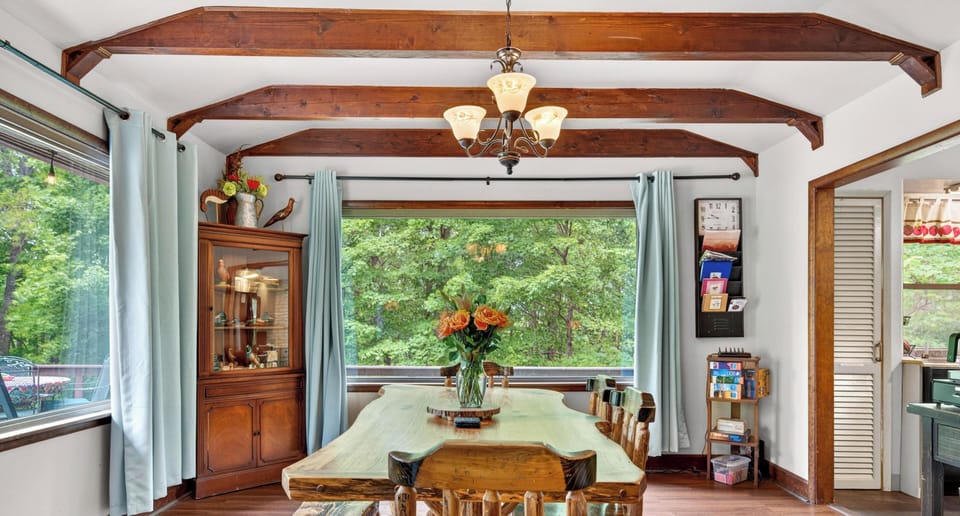 Rustic wood dining table and bench under exposed beams, framed by large windows with blue curtains.
