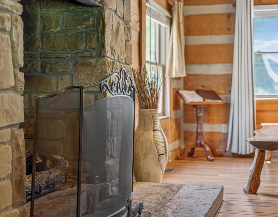 Stone fireplace with wrought iron screen anchors the open dining area, where a rustic wood table and bench sit by bright windows.