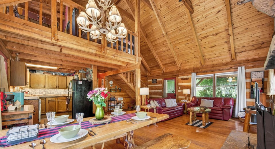 Hand-hewn wood dining table with bench seating faces red leather sofas and lofted ceiling in open living area.