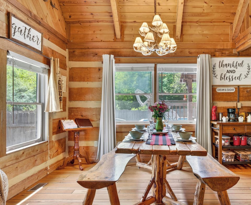 Handcrafted log dining table and benches set with dishes sit beneath antler chandelier and large windows.