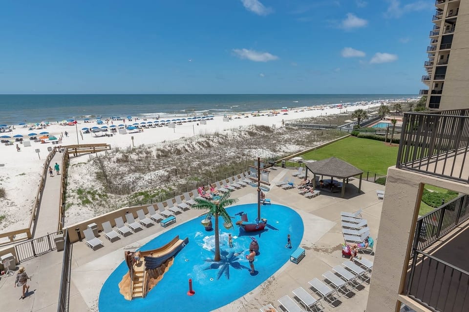 The vibrant pool area features a playful pirate ship splash park and palm trees, easy access to the beach, and sun loungers