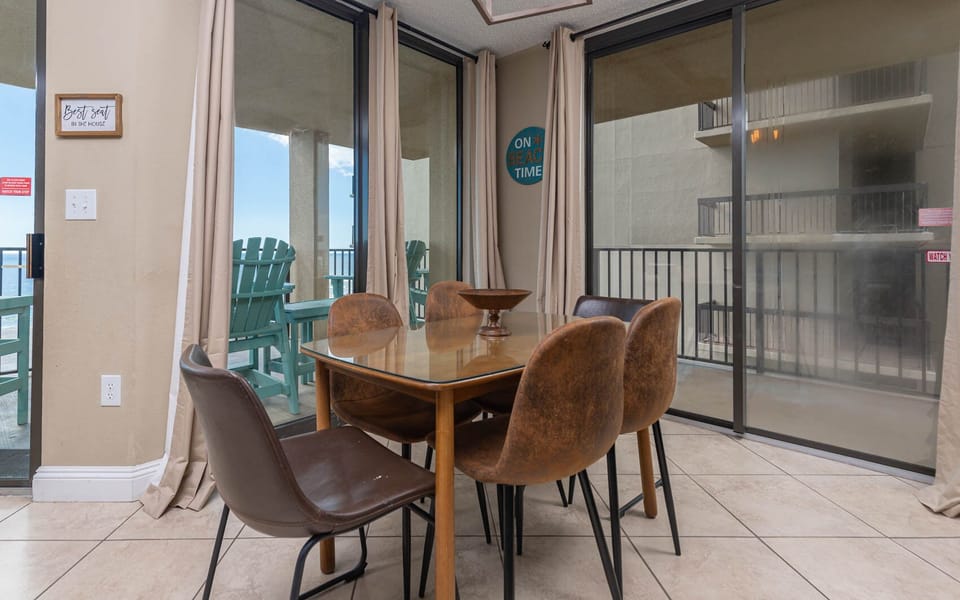 The dining area features a glass-top table surrounded by stylish brown chairs, leading to a beach view balcony