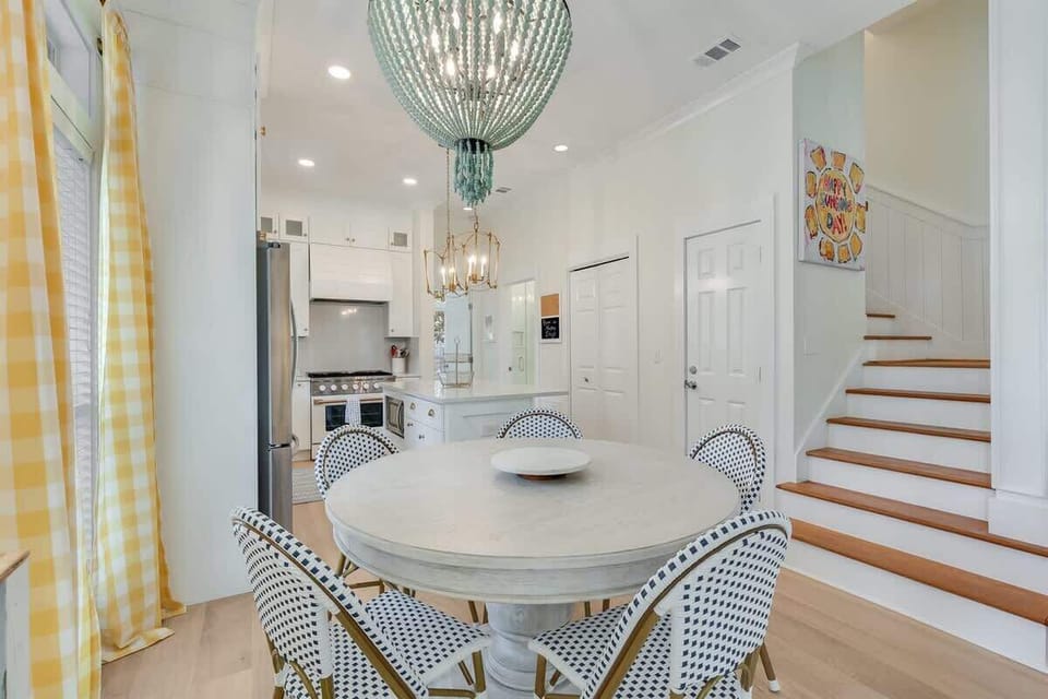 - Round dining table set under a bold coastal chandelier, perfect for family breakfasts or game night - Bright yellow gingham curtains add cheer and charm to the space - Easy flow from kitchen to stairs keeps the layout functional and inviting