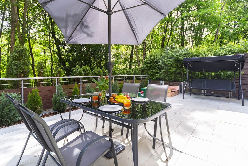 An outdoor dining area with a patio table, four chairs, and a large umbrella. The space is surrounded by greenery, creating a relaxing atmosphere.