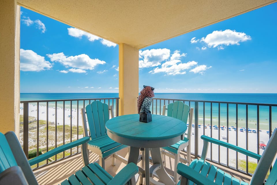 The balcony features a round table with turquoise chairs, offering a picturesque view of the beach and vibrant blue waters