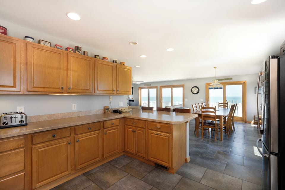 Your fully equipped kitchen flows seamlessly into the dining area, where warm wood tones meet stunning waterfront views through expansive windows—perfect for preparing meals while soaking in the scenery.